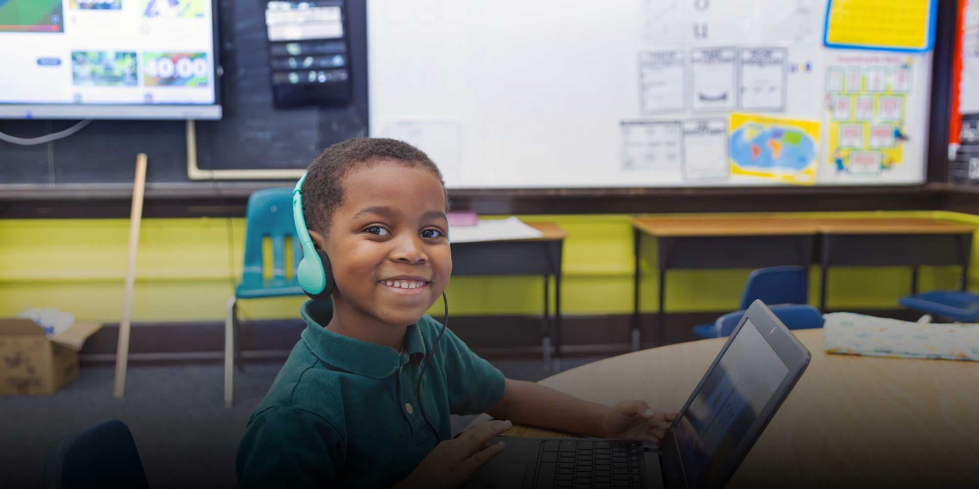 Smiling student wearing headphones at a laptop in a classroom.