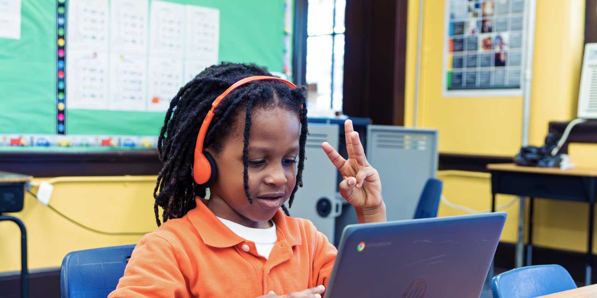 Student in orange headphones holding up two fingers while using a laptop.
